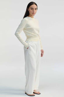 Woman wearing a cream long-sleeve top and white pants on a light gray background
