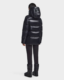 A person faces away, wearing the Rudsak RITA S - L123527 Black womens down puffer jacket with a hood, paired with black pants and black winter boots against a plain light background.