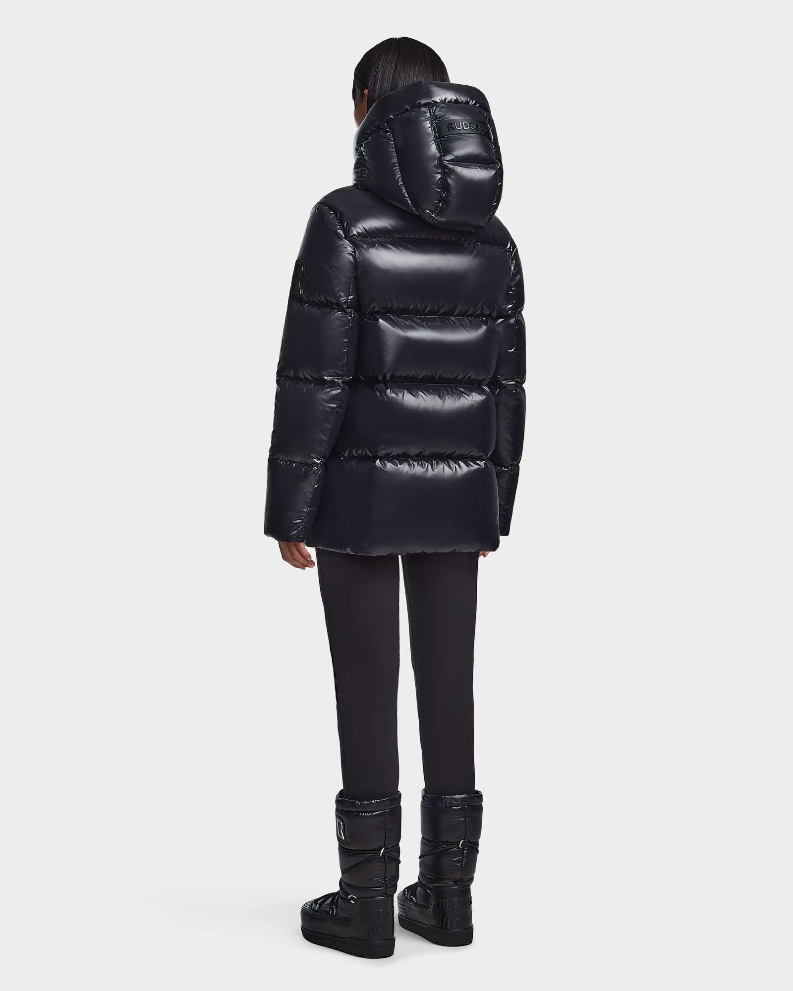A person faces away, wearing the Rudsak RITA S - L123527 Black womens down puffer jacket with a hood, paired with black pants and black winter boots against a plain light background.