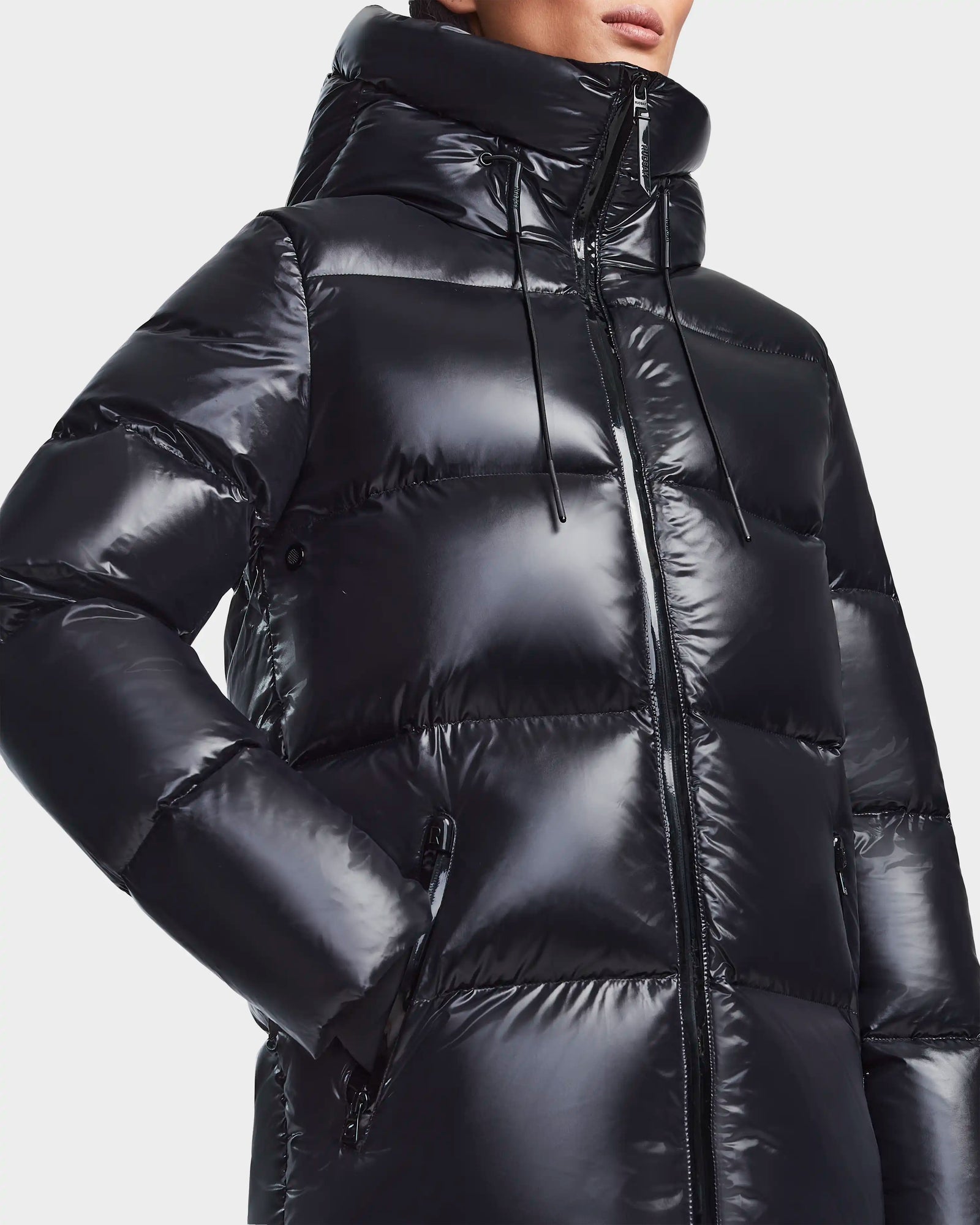 A person wearing the Rudsak RITA S - L123527 Black womens down puffer jacket with a hood and front zipper, featuring large quilted sections, side pockets, and weather-resistant fabric, stands against a plain light background.