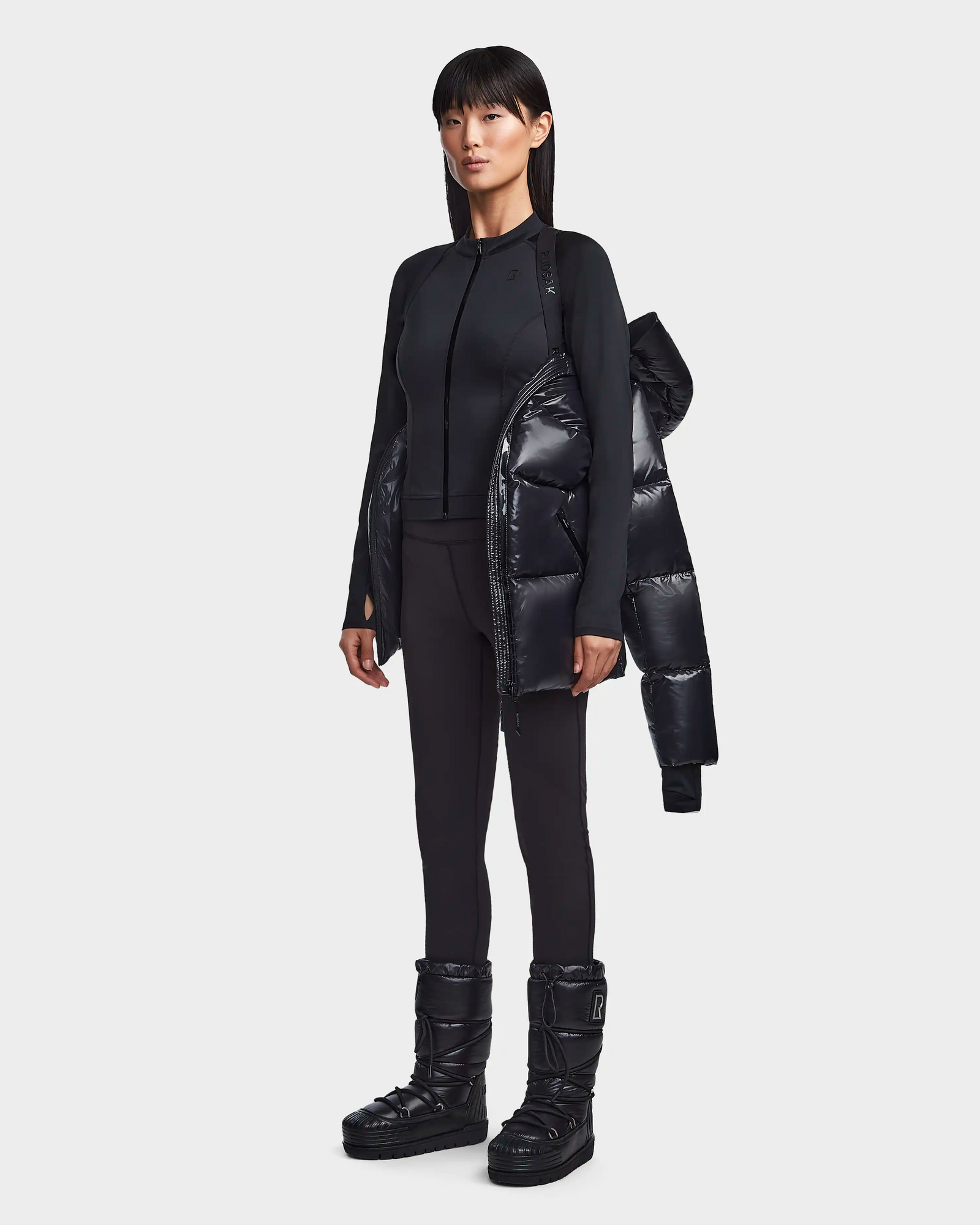 A woman faces forward, wearing the Rudsak RITA S - L123527 Black fitted zip-up jacket, black leggings, weather-resistant puffer boots, and holding a womens down puffer coat; the background is plain and light gray.
