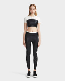 A woman faces forward, wearing the Rudsak TAYLA - 8524015 Black high-performance activewear leggings with white accents and text, a black and white crop top, and black sneakers, set against a plain light gray background.