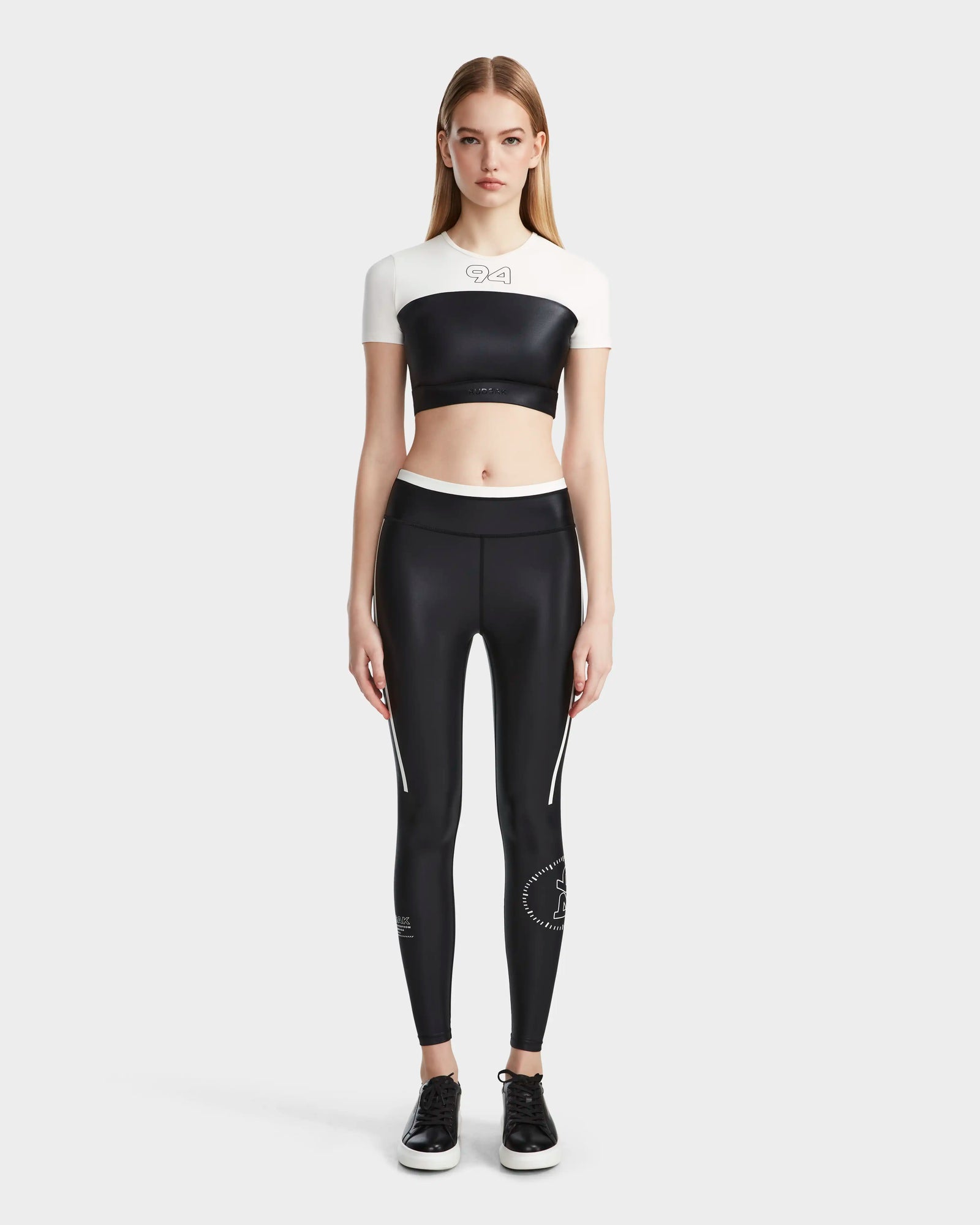 A woman faces forward, wearing the Rudsak TAYLA - 8524015 Black high-performance activewear leggings with white accents and text, a black and white crop top, and black sneakers, set against a plain light gray background.