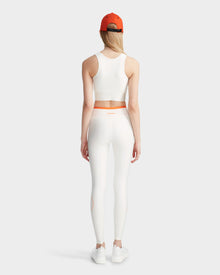 A woman stands facing away, wearing the Rudsak TRACY - 8524016 Eggshell racer-back sports bra and matching leggings, with an orange waistband, white sneakers, an orange cap, and long blonde hair against a light gray background.