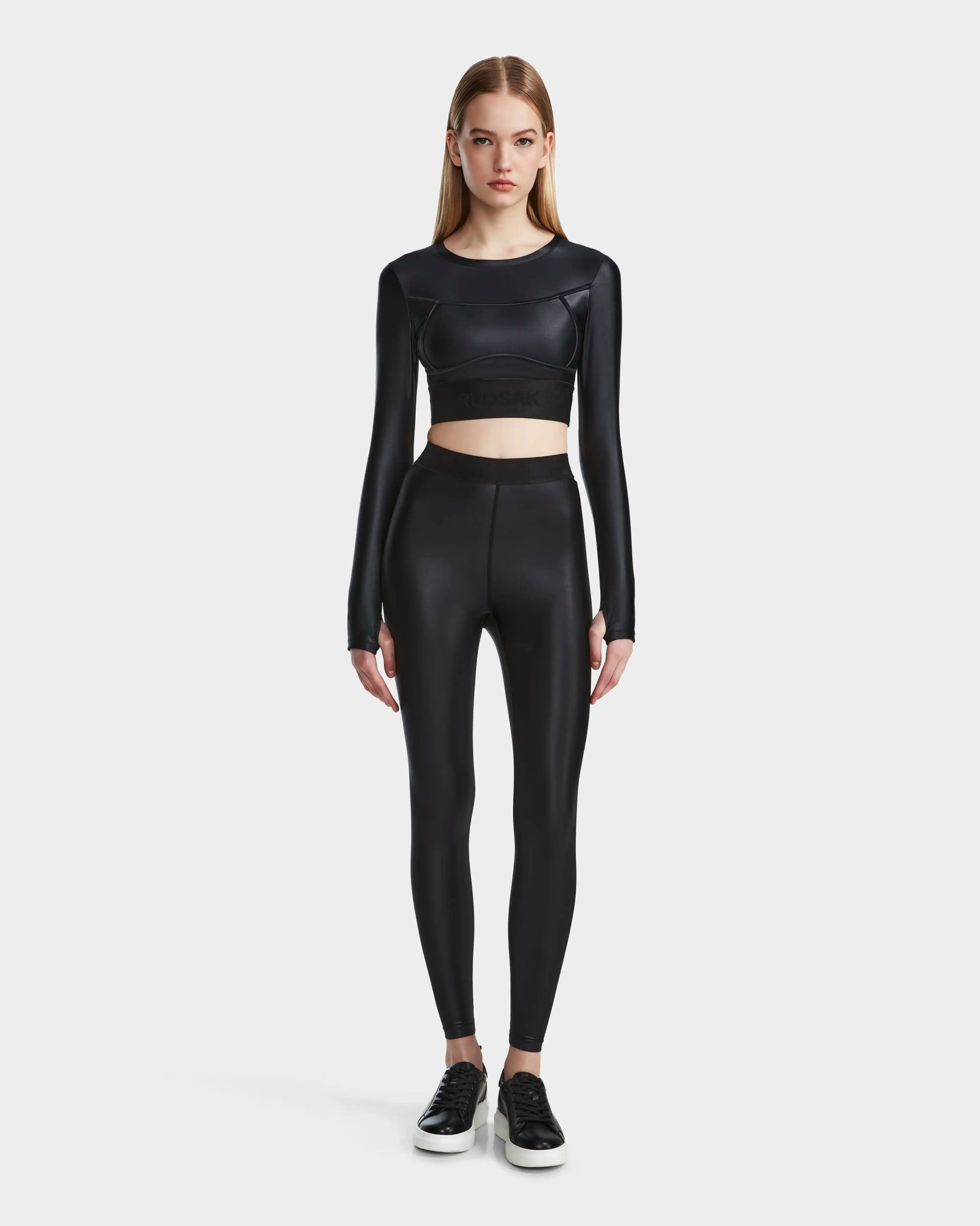 A woman faces forward wearing the Rudsak LORA - 8524026 Black fitted long sleeve top with high-waisted black leggings and sneakers, set against a plain light background.