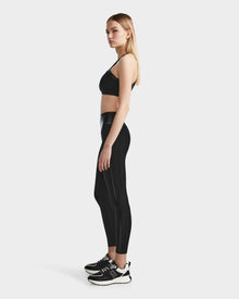 A woman with straight blonde hair poses in profile against a white background, dressed in Rudsaks THEA - 8523003 BLACK high-rise leggings with a vegan leather waistband, styled with a black sports bra and sneakers.