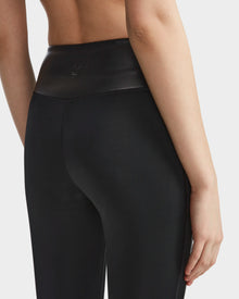 A person models the Rudsak THEA - 8523003 BLACK high-rise leggings with a shiny vegan leather waistband, viewed from the back and waist down. Their right arm rests by their side against a plain white background.