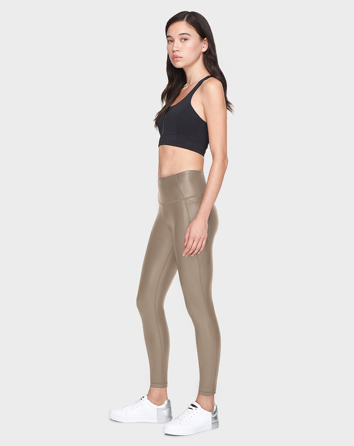 A woman stands in profile against a plain background, wearing the Rudsak SHIRIN L - 8521034 Cement leggings and a black sports bra with white sneakers. She has long, dark hair and a neutral expression.