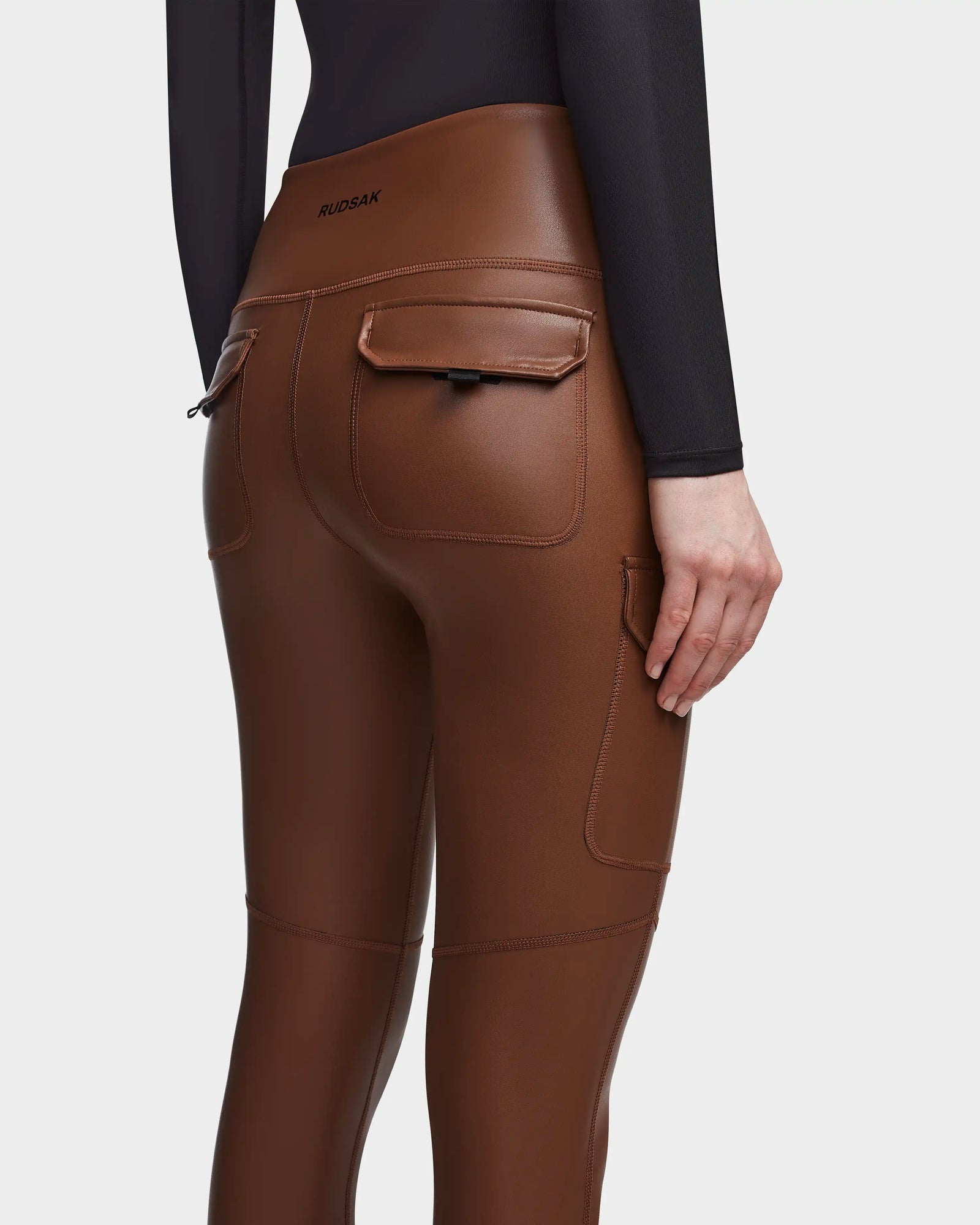 A person wears Rudsak SIEN L - 8521042 COHIBA brown vegan leather leggings with back pockets and a black long-sleeve top, shown from the waist down. The Rudsak logo appears on the waistband—perfect activewear essentials.