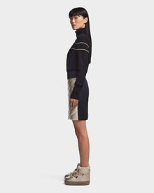 A woman stands in profile against a plain background, wearing the MARZI - 8523528 Metallic Ivory winter boots from Rudsak, styled with a black and white striped turtleneck sweater and a beige and black chevron quilted skirt.