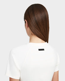 A woman with straight brown hair, seen from behind, wears the Rudsak SUMI - 8524030 Cotton White essential tee with fitted short sleeves and a small black rectangular label on the upper back. The background is plain light gray.