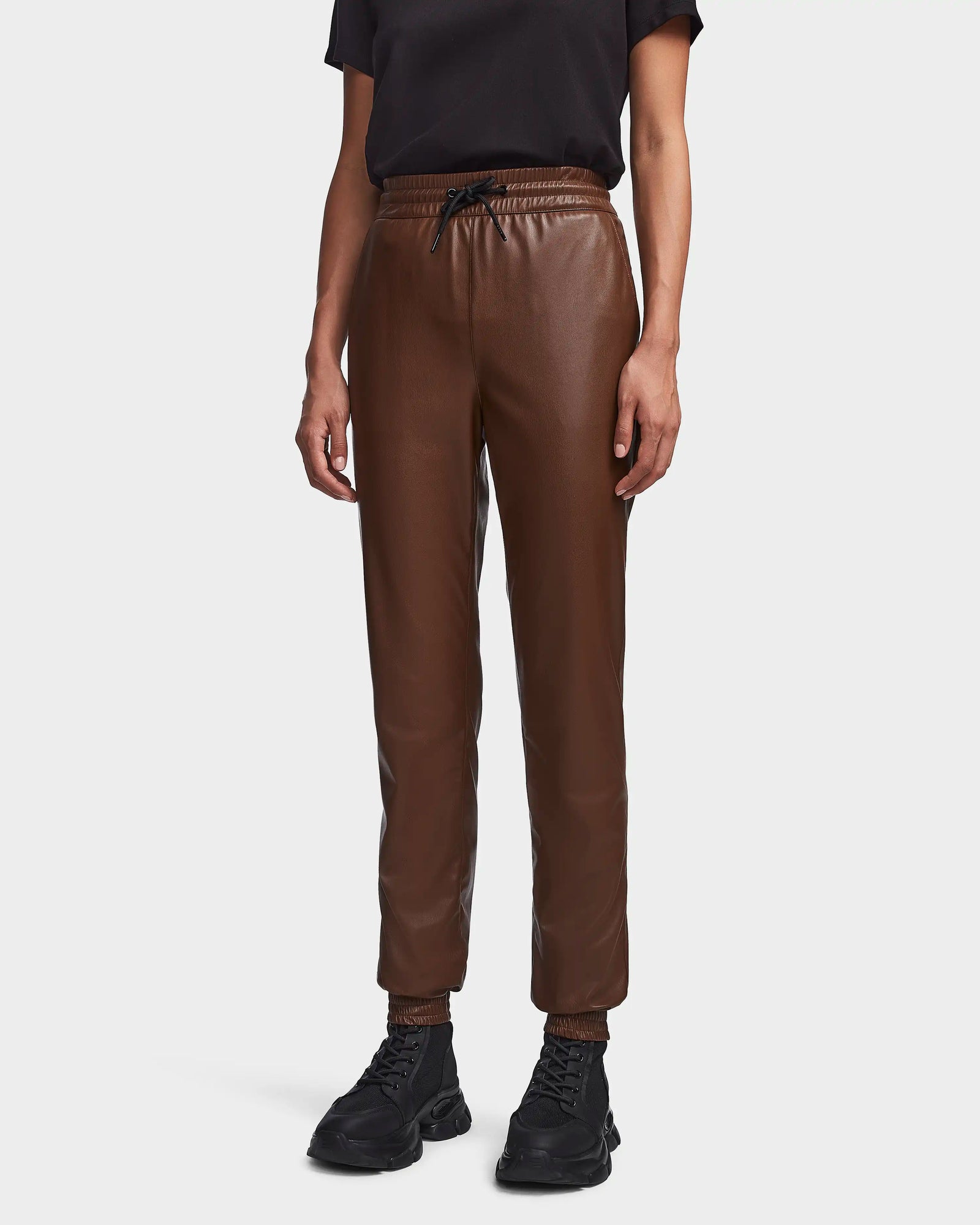 A woman models Rudsak's FELICIA - 8523025 COHIBA brown vegan leather joggers, featuring a visible drawstring and elastic waistband, paired with a black short-sleeve shirt and black sneakers against a plain white background.