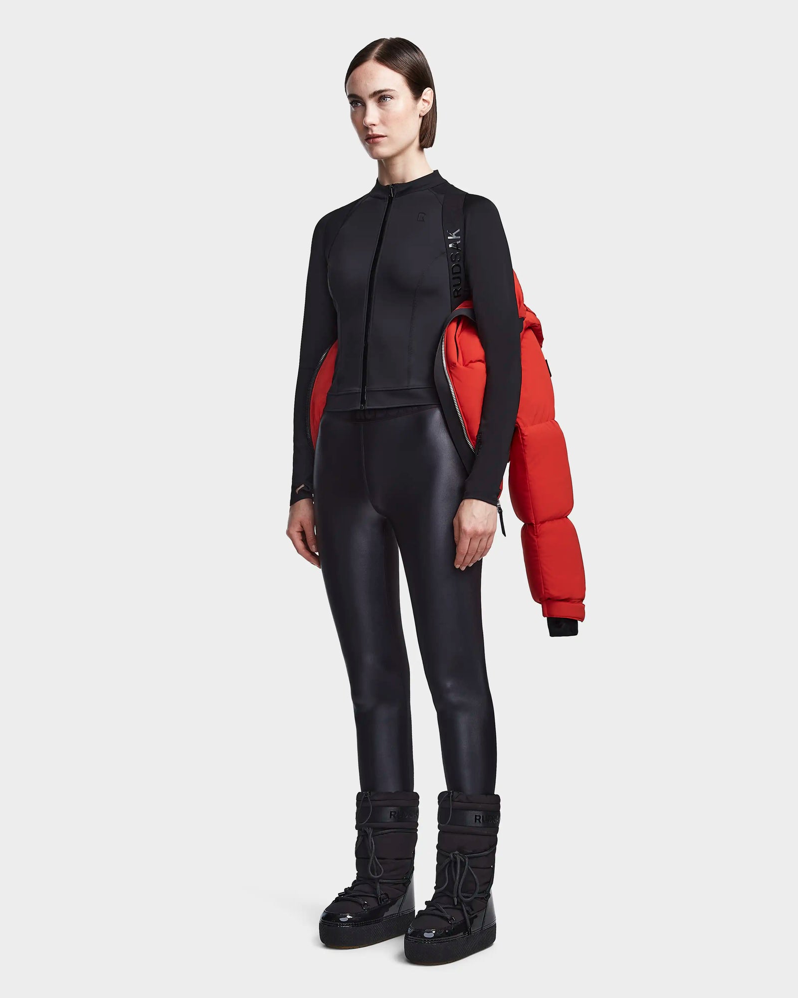 A woman wears a fitted black zip-up jacket, matching leggings, and black winter boots, carrying the Rudsak MAVE M - 8123572 Orange puffer over her shoulder against a plain white background.