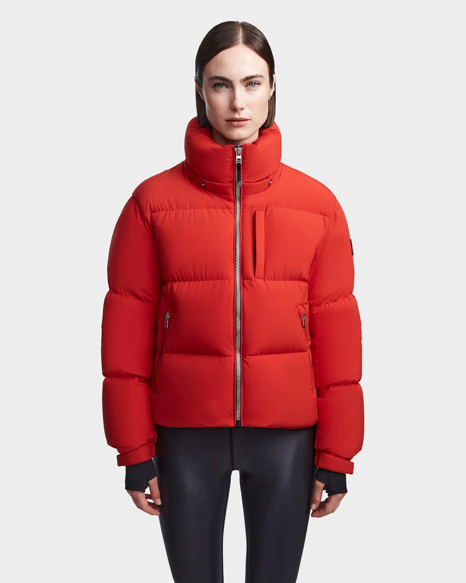 A woman with straight brown hair models the Rudsak MAVE M - 8123572 Orange down puffer jacket with a high collar and front zipper, paired with black leggings, against a plain light gray background.