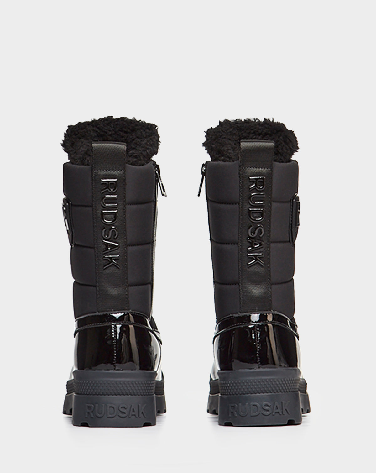 A pair of waterproof Rudsak LEX - 8222501 BLACK women’s boots with quilted sides, patent leather heels, and thick rugged soles, shown from behind against a white background.