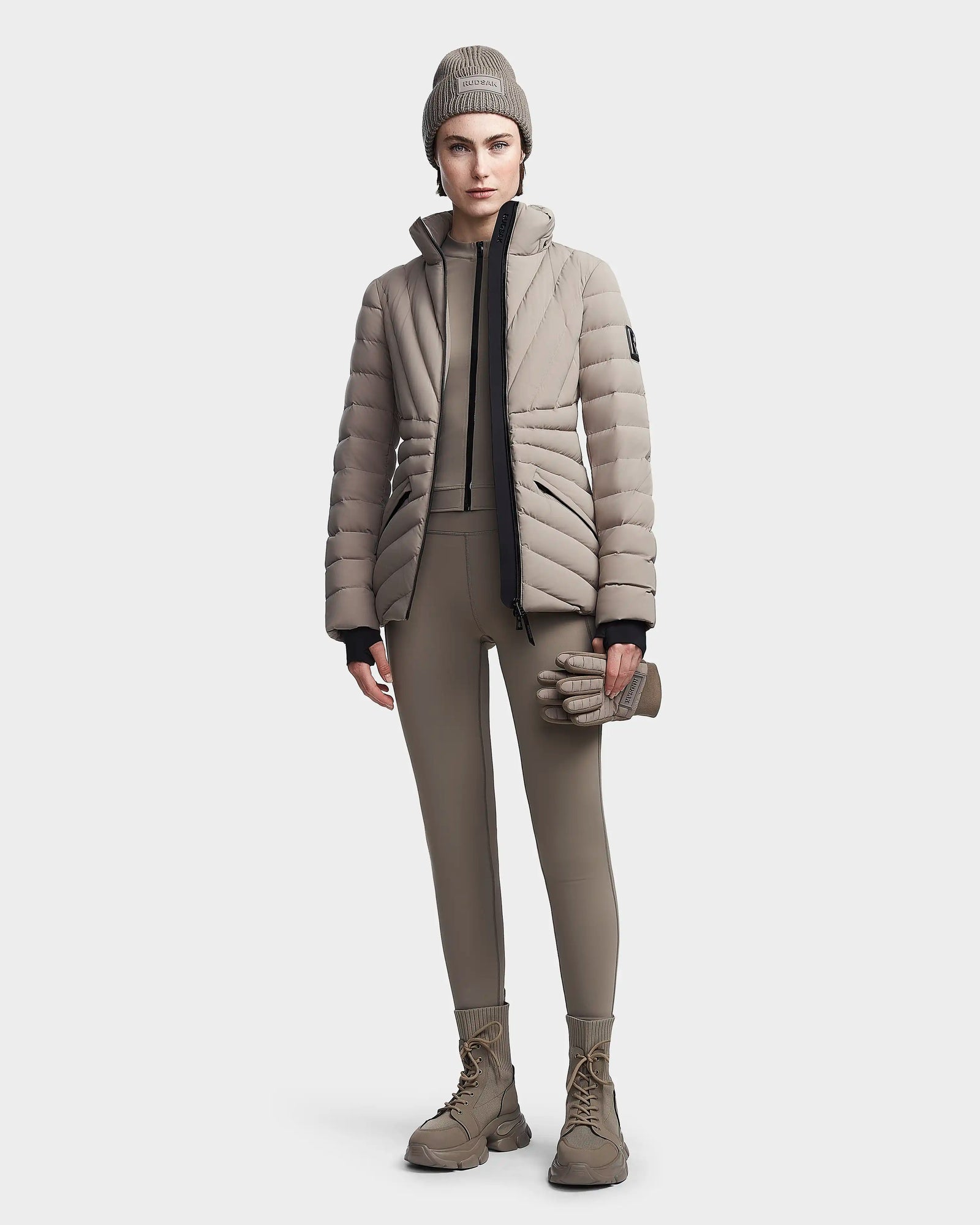 A woman faces forward in a modern monochromatic look, wearing the Rudsak HANNA - 8123562 Cement RDS certified winter down jacket, matching beanie, tan leggings, lace-up boots, and holding tan gloves against a neutral backdrop.