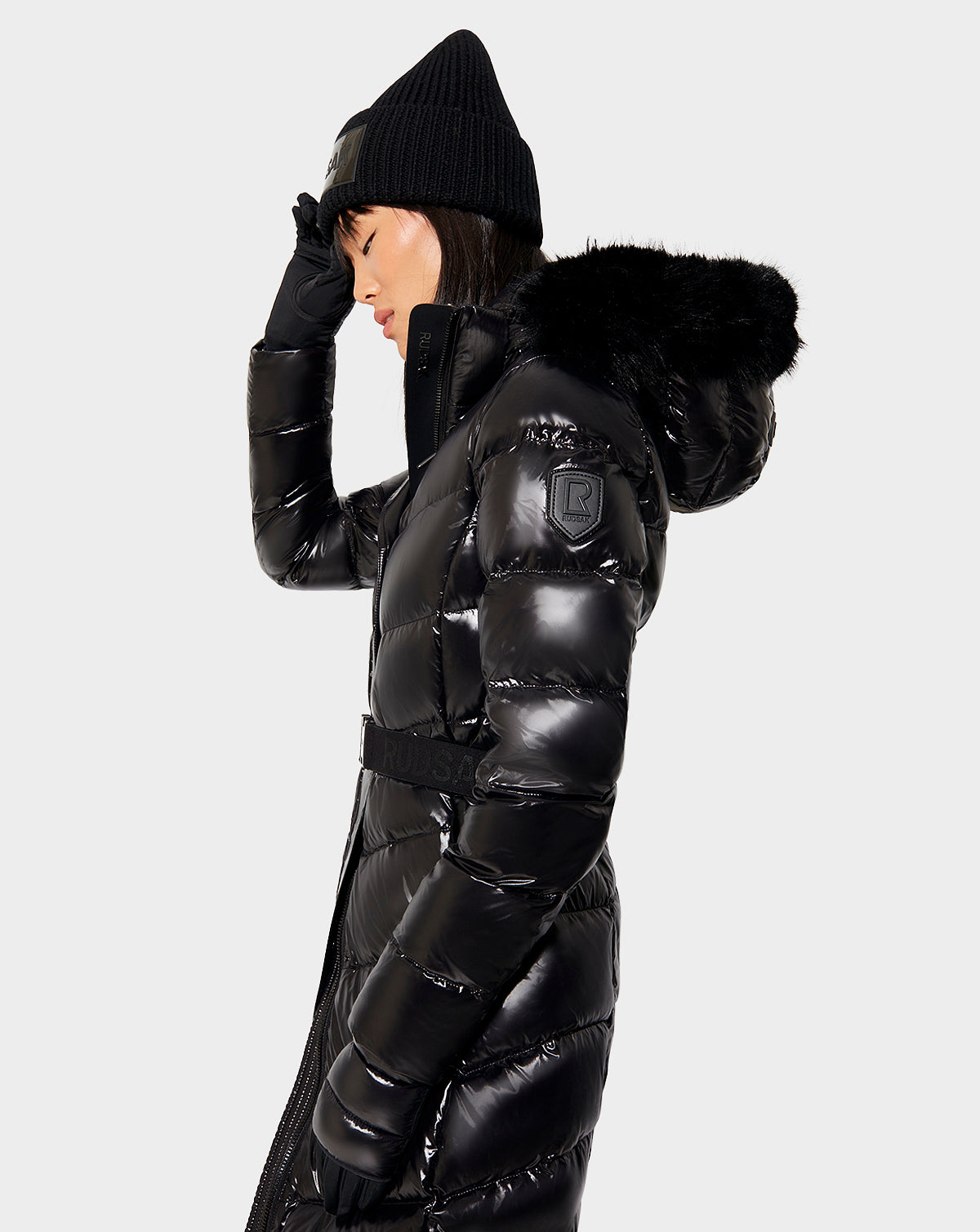A woman wearing the Rudsak ELSIE - L121525 BLACK shiny black maxi down puffer coat with removable hood, plus a black knit hat and gloves, stands in profile against a plain light background, touching her hat with one hand.