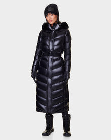 A woman wearing the Rudsak ELSIE - L121525 BLACK maxi down puffer coat with a belted waist, fur-lined hood, black beanie, gloves, and lace-up black boots stands against a plain light background.