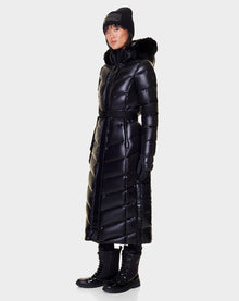 A person wears the Rudsak ELSIE - L121525 BLACK women’s maxi down puffer with RDS-certified down and a fur-trimmed hood, paired with black gloves, winter boots, and a black beanie, standing against a plain light background.