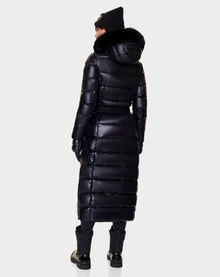 A person is seen from the back wearing the Rudsak ELSIE - L121525 BLACK women’s maxi down puffer with a recycled semi-gloss exterior, fur-trimmed hood, and shiny black finish, paired with black gloves, pants, shoes, and knit hat against a light background.