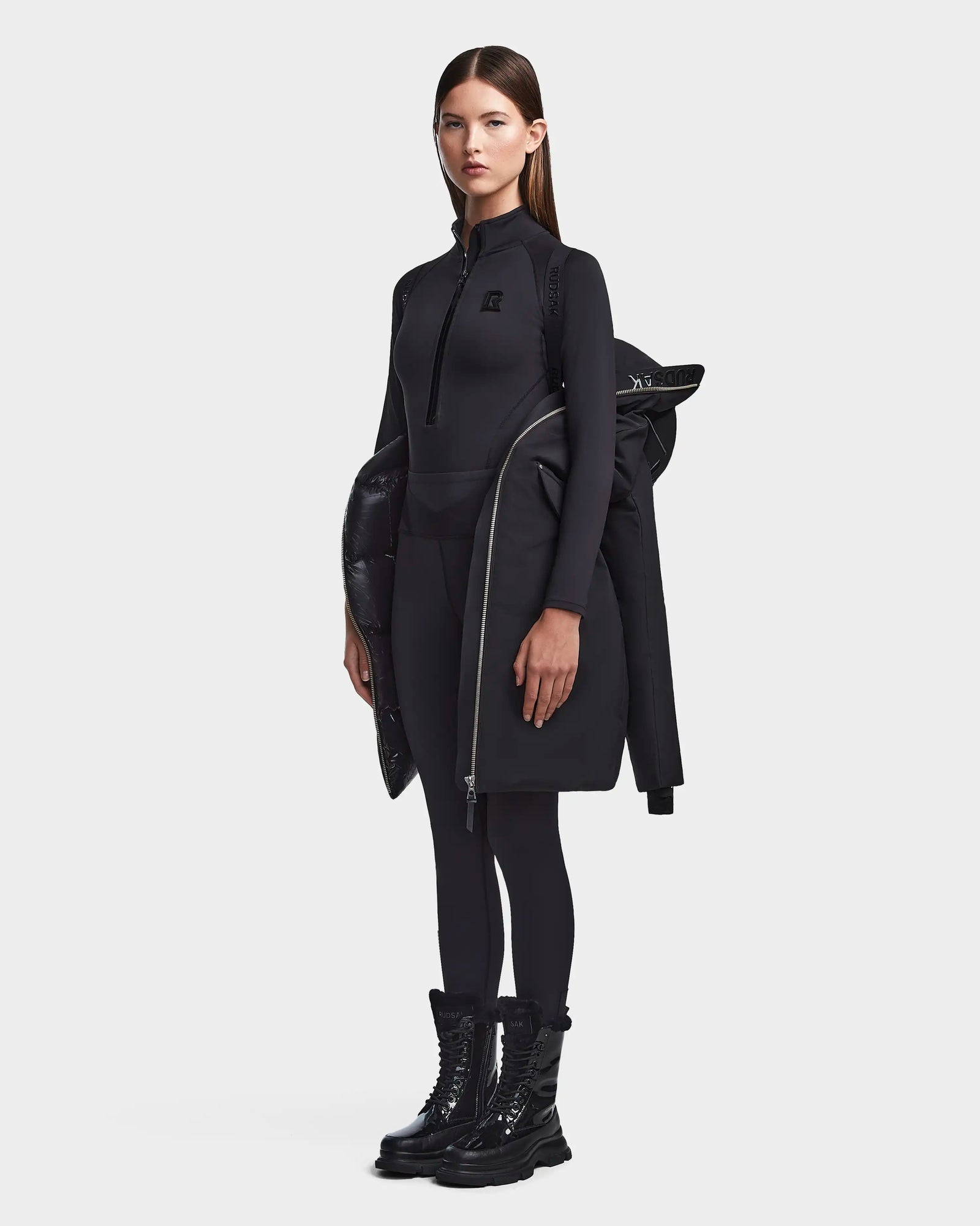 A woman stands against a light background wearing the RUDSAK MILENA - 8122536 BLACK womens winter coat, with a zipper top, black leggings, chunky boots, and the coat made from recycled Stormshell fabric draped over one shoulder.