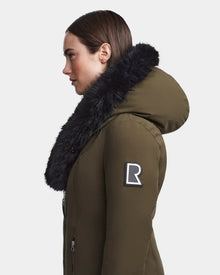 A woman in profile wears the Rudsak MAIA M - 8123582 Army down parka with a large black fur collar and hood. A bold R patch accents the upper sleeve. The plain white background highlights the olive green color.