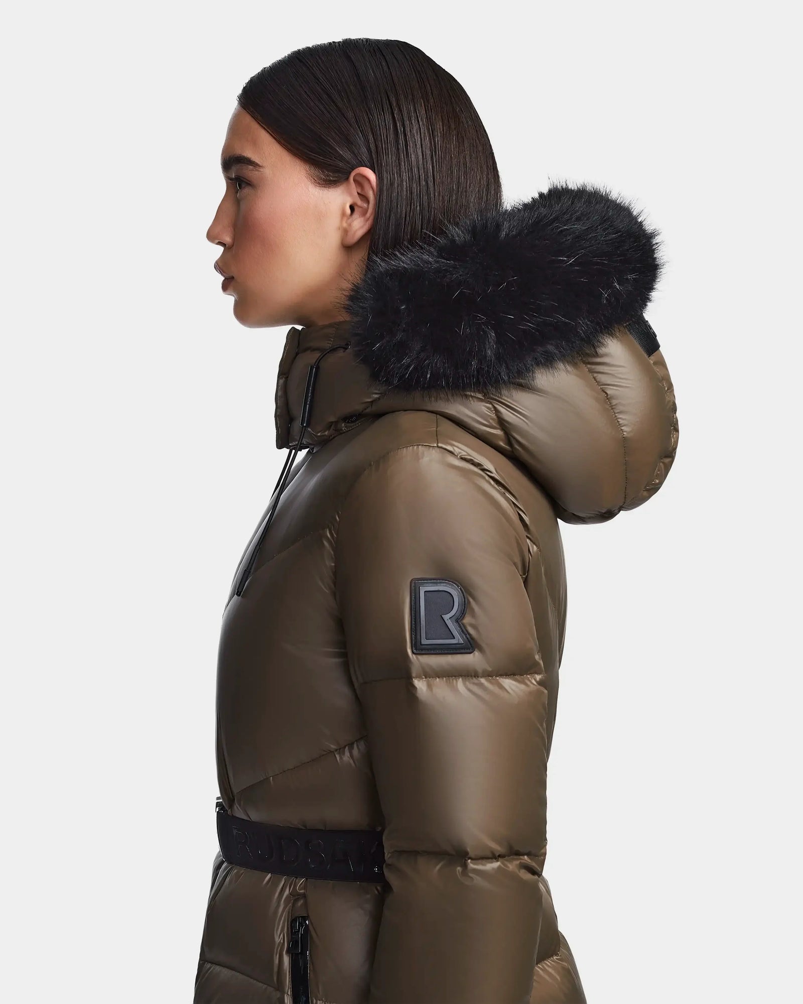 A person in profile models the Rudsak CHRISTELLE S - L123524 Light Olive women's chevron quilted down puffer with a black fur-trimmed hood, sleeve R patch, waist belt, set against a plain white background.