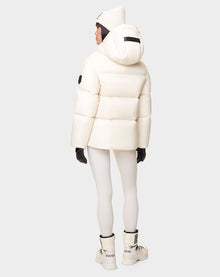 A person stands facing away, wearing the RUDSAK DORA - 8122547 ICELAND womens white down-filled puffer jacket with removable hood, white leggings, winter boots, black gloves, and a knit beanie against a plain light background.