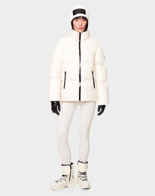 A woman wears the RUDSAK DORA - 8122547 ICELAND white down-filled puffer jacket with removable hood, white leggings, boots, black gloves, and a white knit hat featuring a black patch, standing against a plain light gray background.