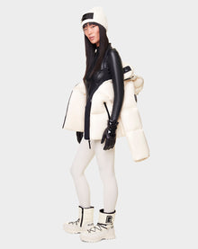 A woman wears the RUDSAK DORA - 8122547 ICELAND down-filled puffer jacket over a black top, white beanie, black gloves, white leggings, and chunky white boots against a plain light gray background.