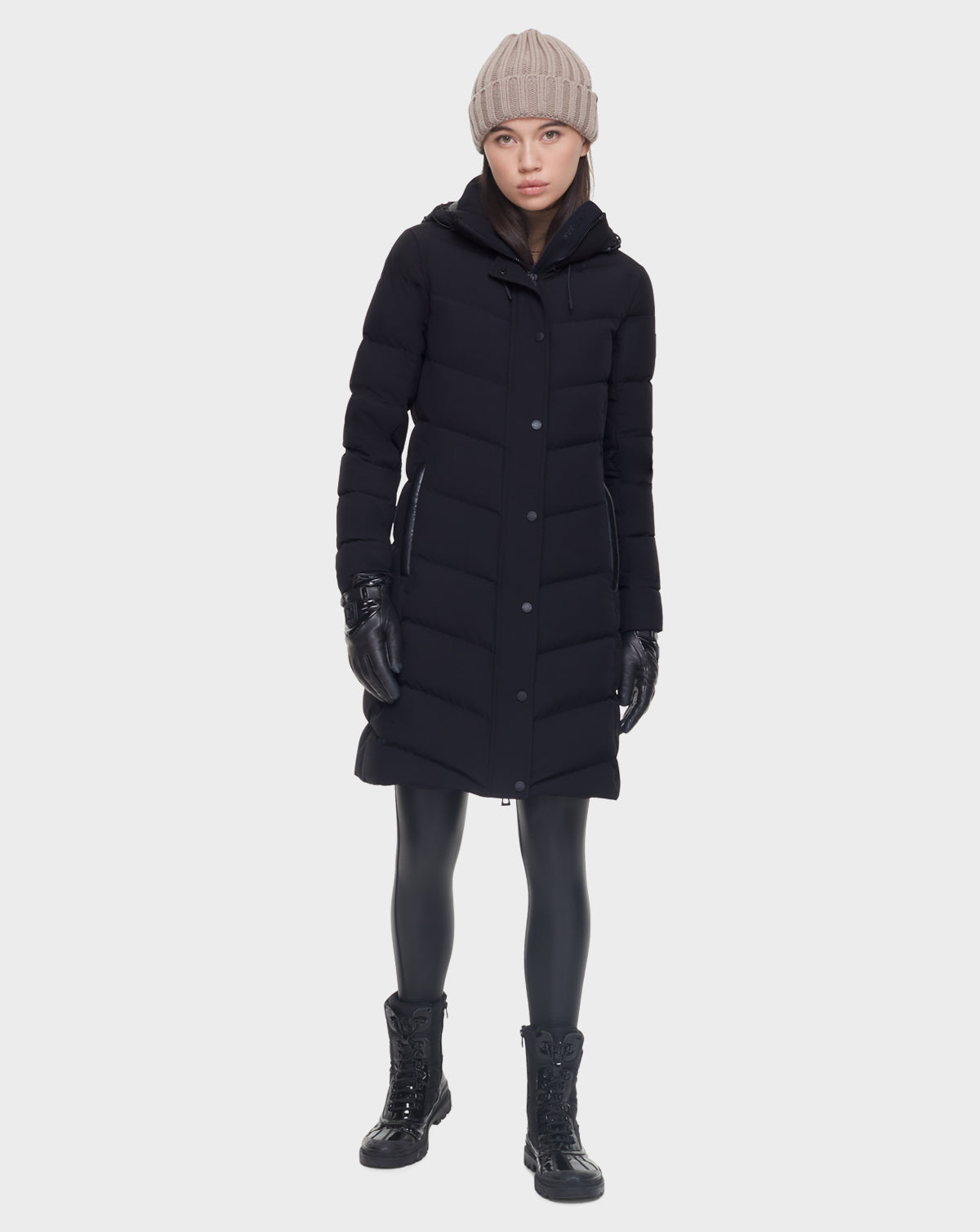A woman stands facing forward against a plain white background wearing the Rudsak ELISA - L121512 BLACK womens quilted down puffer coat, styled with black gloves, leggings, boots, and a beige knit beanie.