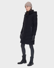 A person wears the Rudsak ELISA - L121512 BLACK womens quilted down puffer with black gloves, leggings, boots, and a beige knit beanie, standing against a plain white background.