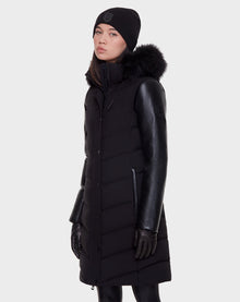 A person wears the Rudsak ELSA - L121519 BLACK sustainable winter coat with leather sleeves and a fur-lined hood, paired with black gloves and a black beanie, standing against a plain light background.