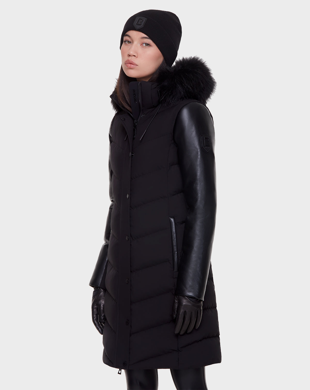 A person wears the Rudsak ELSA - L121519 BLACK sustainable winter coat with leather sleeves and a fur-lined hood, paired with black gloves and a black beanie, standing against a plain light background.