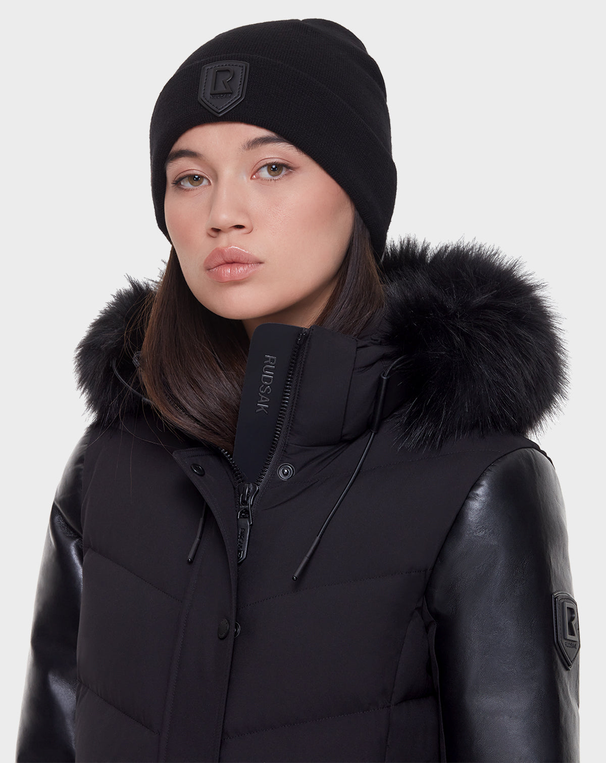 A woman in the Rudsak ELSA - L121519 BLACK sustainable winter coat with leather sleeves and a fur-trimmed hood, and a black beanie, looks at the camera with a neutral expression.