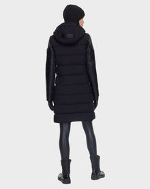 A person wearing the Rudsak ELSA - L121519 BLACK women’s quilted down puffer coat, along with black gloves, pants, and boots, stands facing away from the camera against a plain light background.
