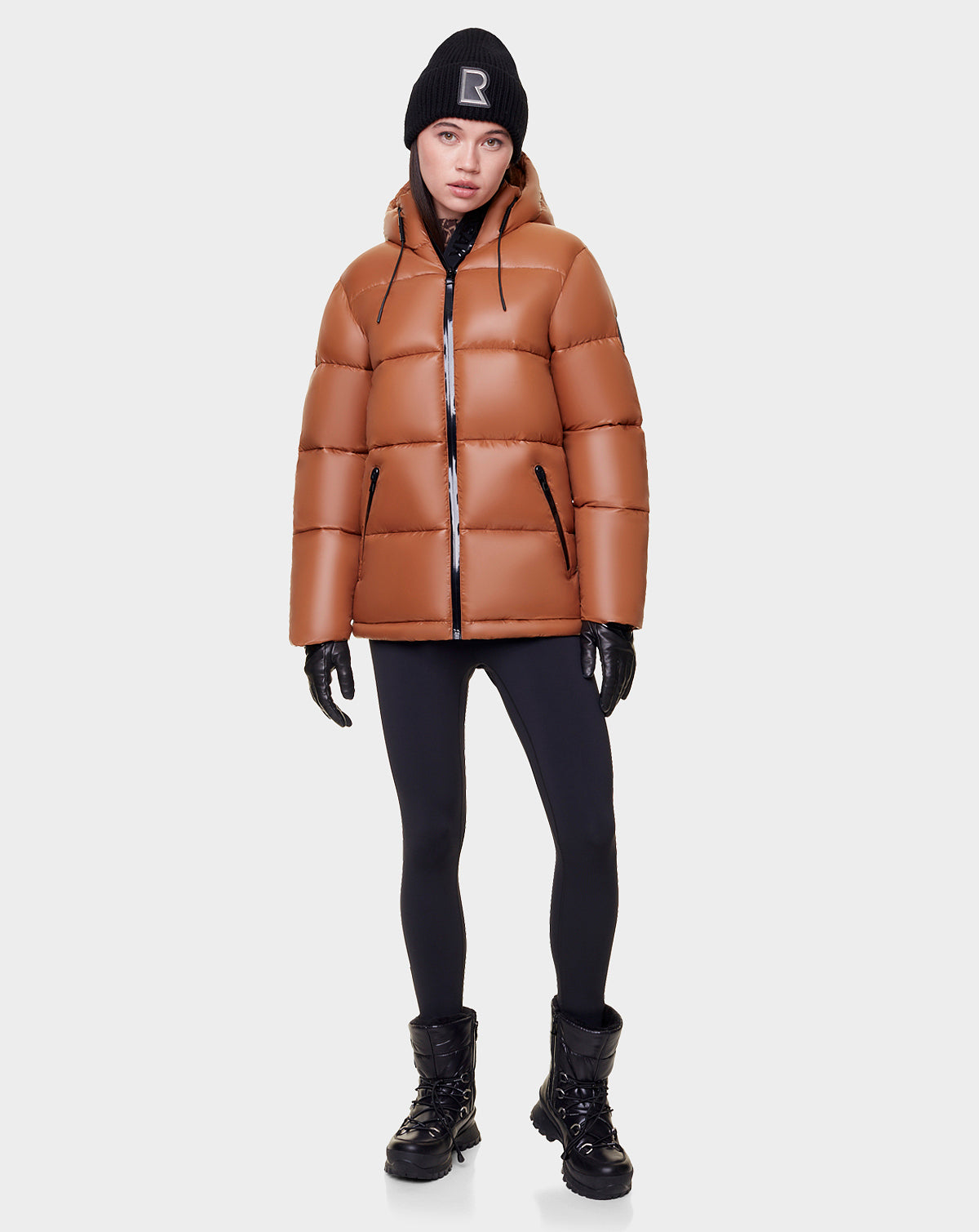 A woman wearing the Rudsak EVIE - L122517 DARK CAMEL puffer, black leggings, gloves, winter boots, and a black beanie with an R stands against a plain white background.