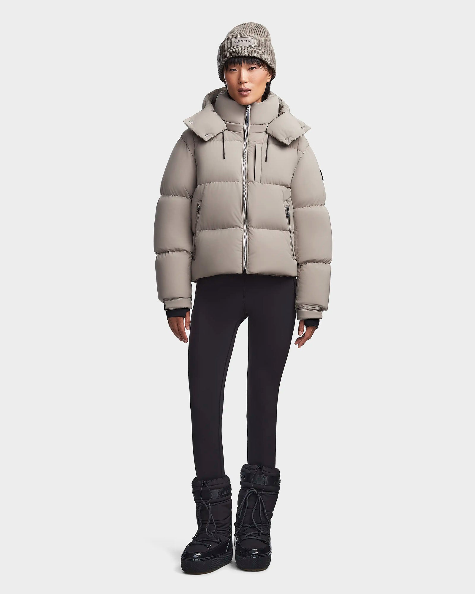 A person wears the Rudsak MAVE M - 8123572 Cement womens down puffer jacket with a matching knit beanie, black leggings, and black winter boots, standing against a plain white background.