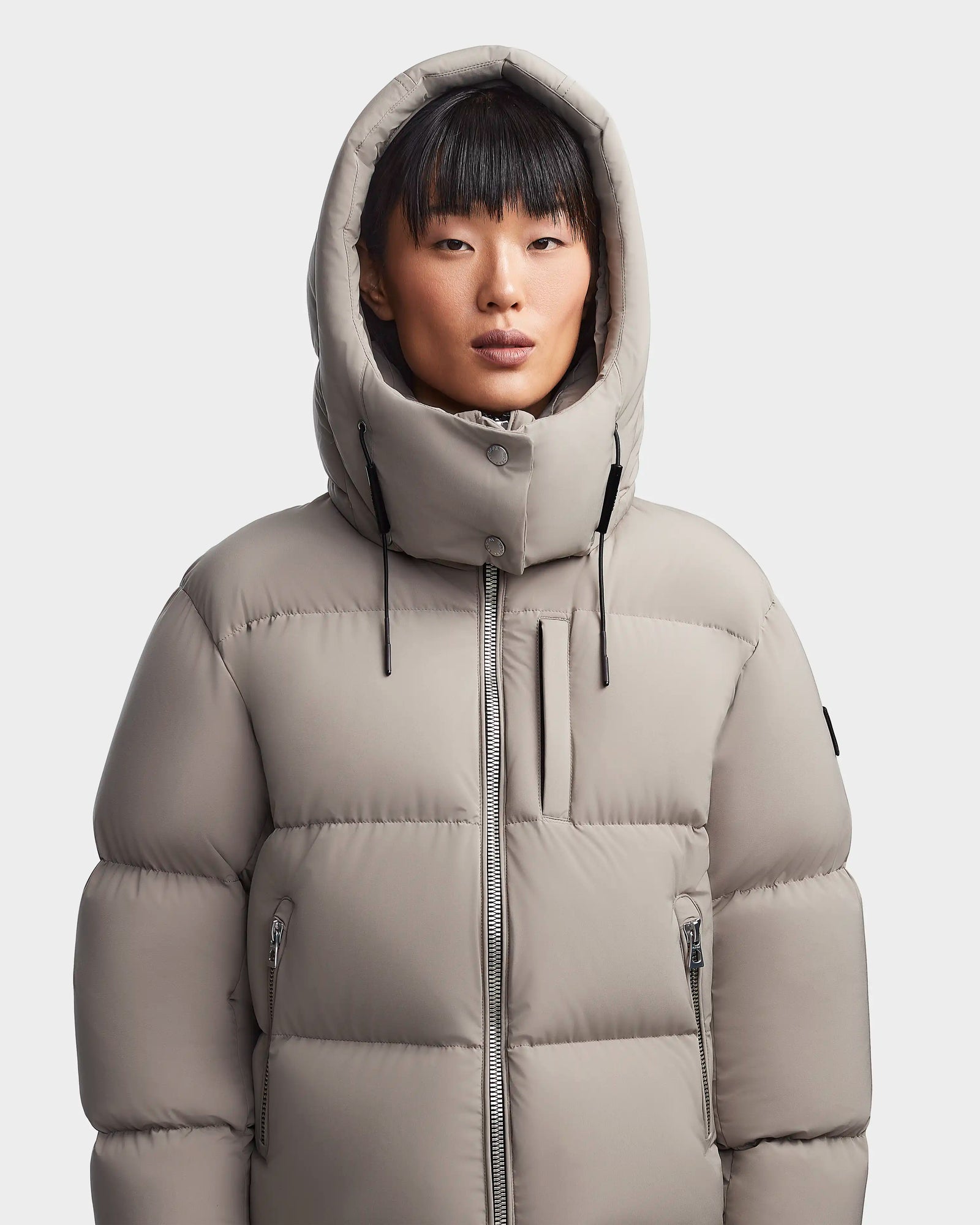 A person with straight black hair and bangs wears the Rudsak MAVE M - 8123572 Cement women’s down puffer jacket with hood up. The water-repellent jacket features zipped pockets and a high collar, standing against a light background.
