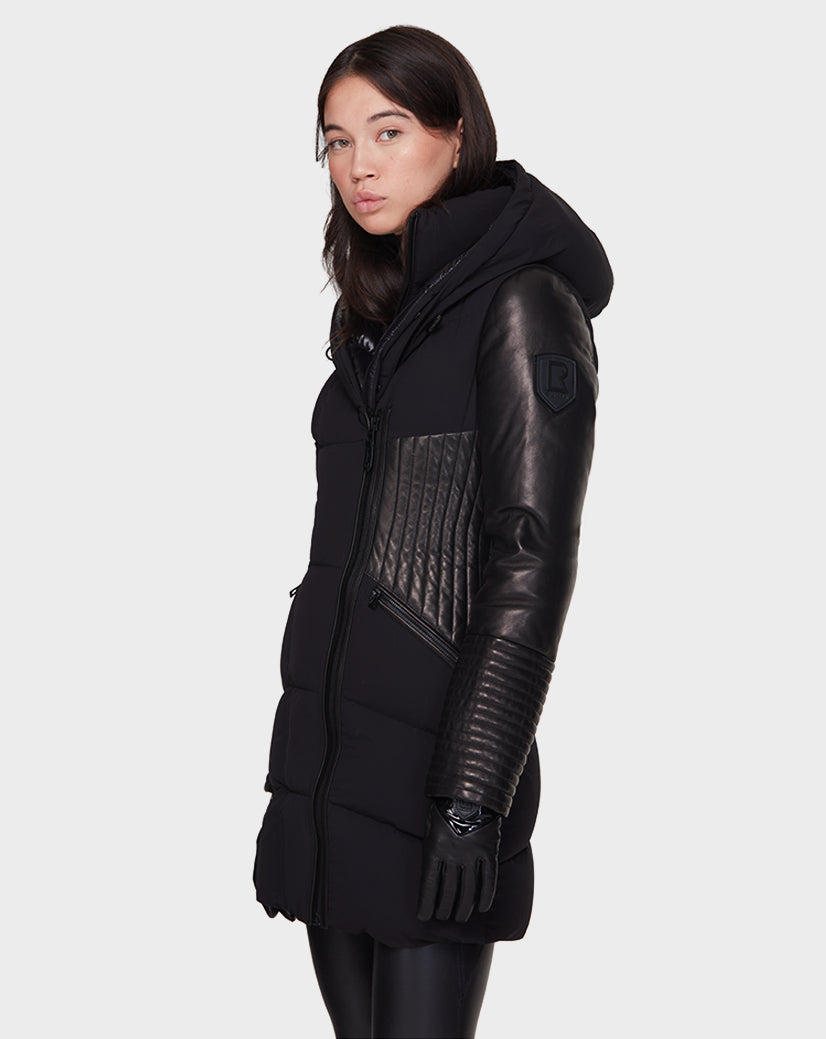A woman wearing the Rudsak SHAUNA - 8121509 BLACK long, hooded winter coat with quilted leather sleeves and side panels stands against a plain light gray background.