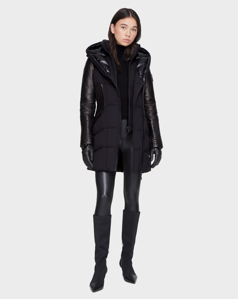 A woman faces forward wearing the Rudsak SHAUNA - 8121509 BLACK quilted down puffer coat with leather sleeves, styled with black gloves, shiny leggings, and knee-high boots against a plain light gray background.