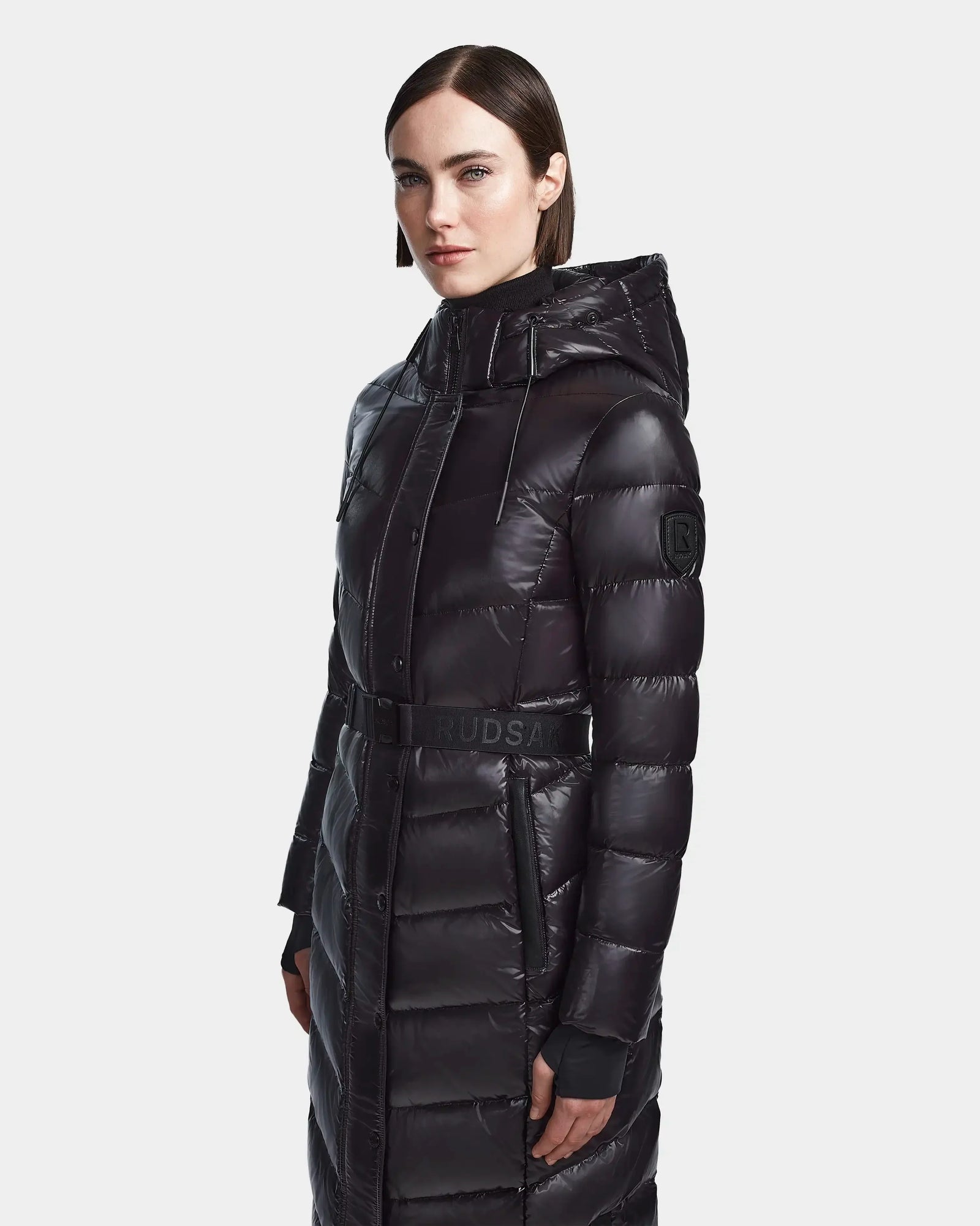 A woman with straight, dark brown hair wears the Rudsak ELSIE S - L123518 Black maxi down puffer coat with a hood and belted waist, standing against a plain light background.