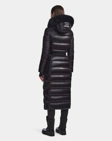 A person stands facing away, wearing the Rudsak ELSIE S - L123518 Black womens maxi down puffer coat with black fur-trimmed hood. The long, RDS-certified down coat extends below the knees, paired with black leggings and boots on a white background.