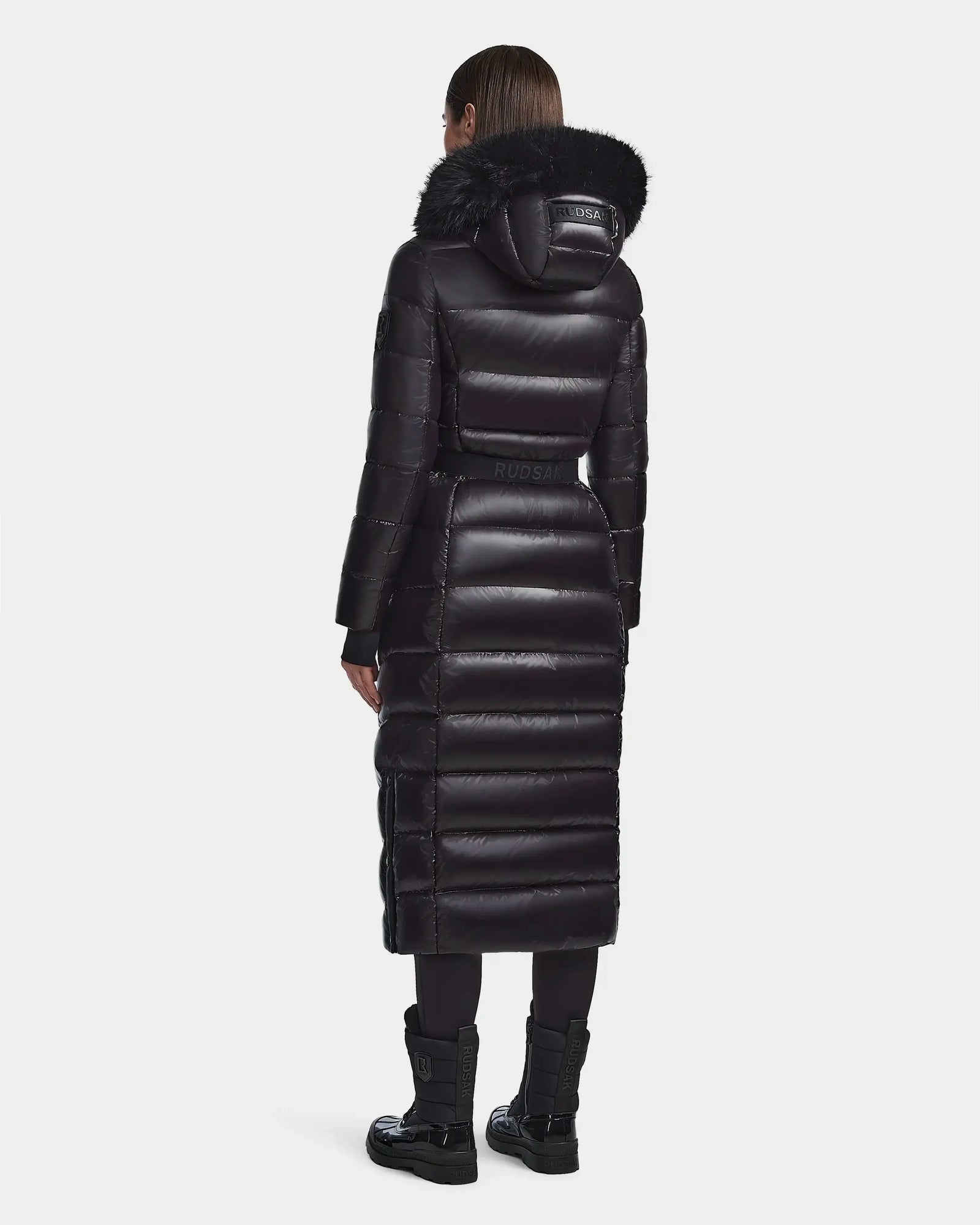 A person stands facing away, wearing the Rudsak ELSIE S - L123518 Black womens maxi down puffer coat with black fur-trimmed hood. The long, RDS-certified down coat extends below the knees, paired with black leggings and boots on a white background.