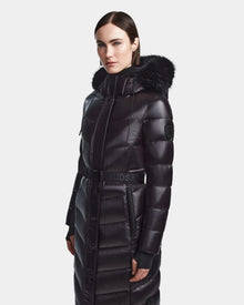 A woman wears the Rudsak ELSIE S - L123518 Black maxi down puffer with a belted waist and fur-trimmed hood, standing against a plain light gray background.