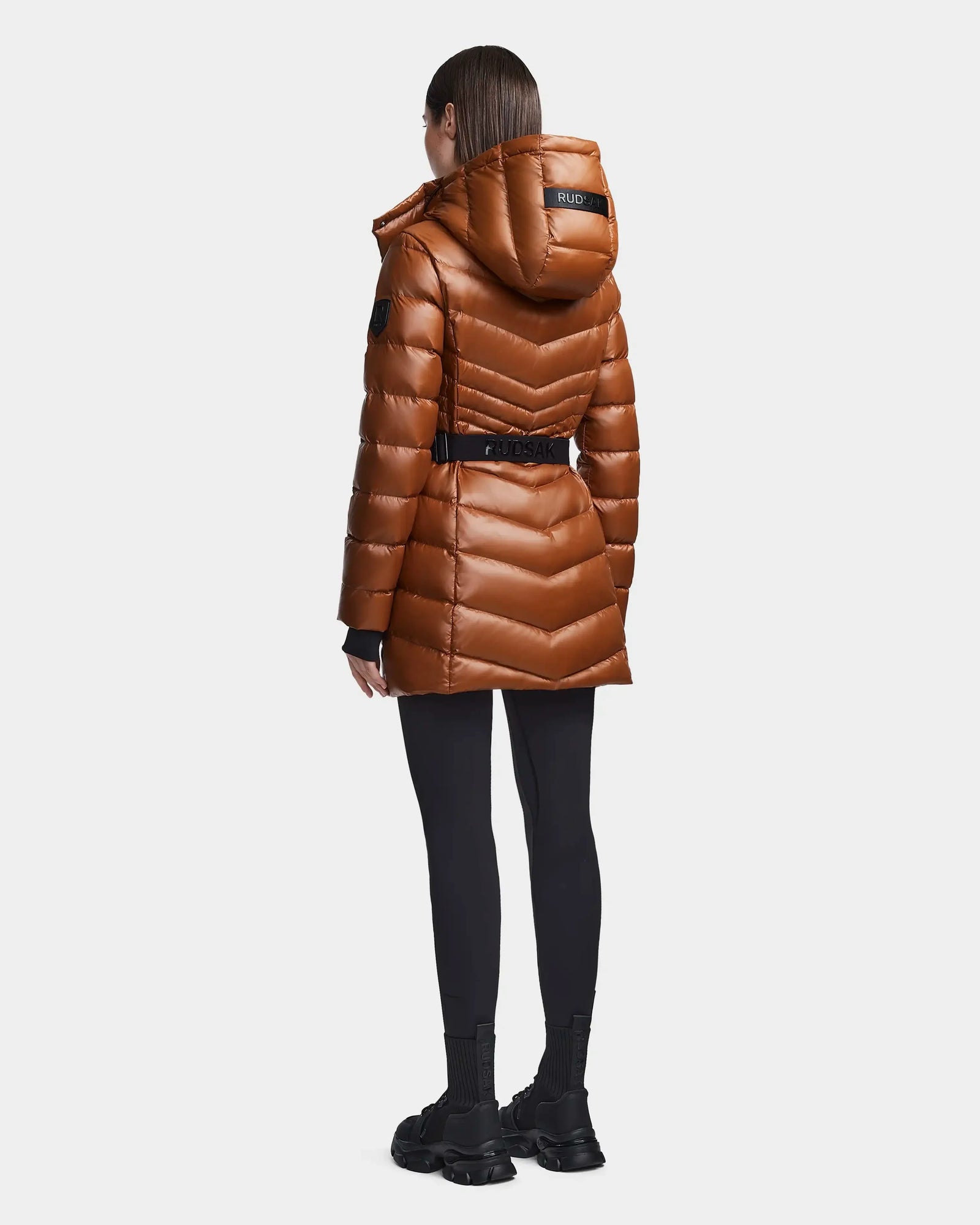 A person with straight brown hair wears the Rudsak MALAYA - L123510 Dark Camel women’s down puffer jacket with a belt, black leggings, and chunky black boots, standing facing away against a light background.