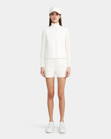 A woman faces forward wearing the Rudsak LOUISE - 8124046 Eggshell lightweight quilted jacket with matching white shorts, styled with a white baseball cap and sneakers against a plain light background.