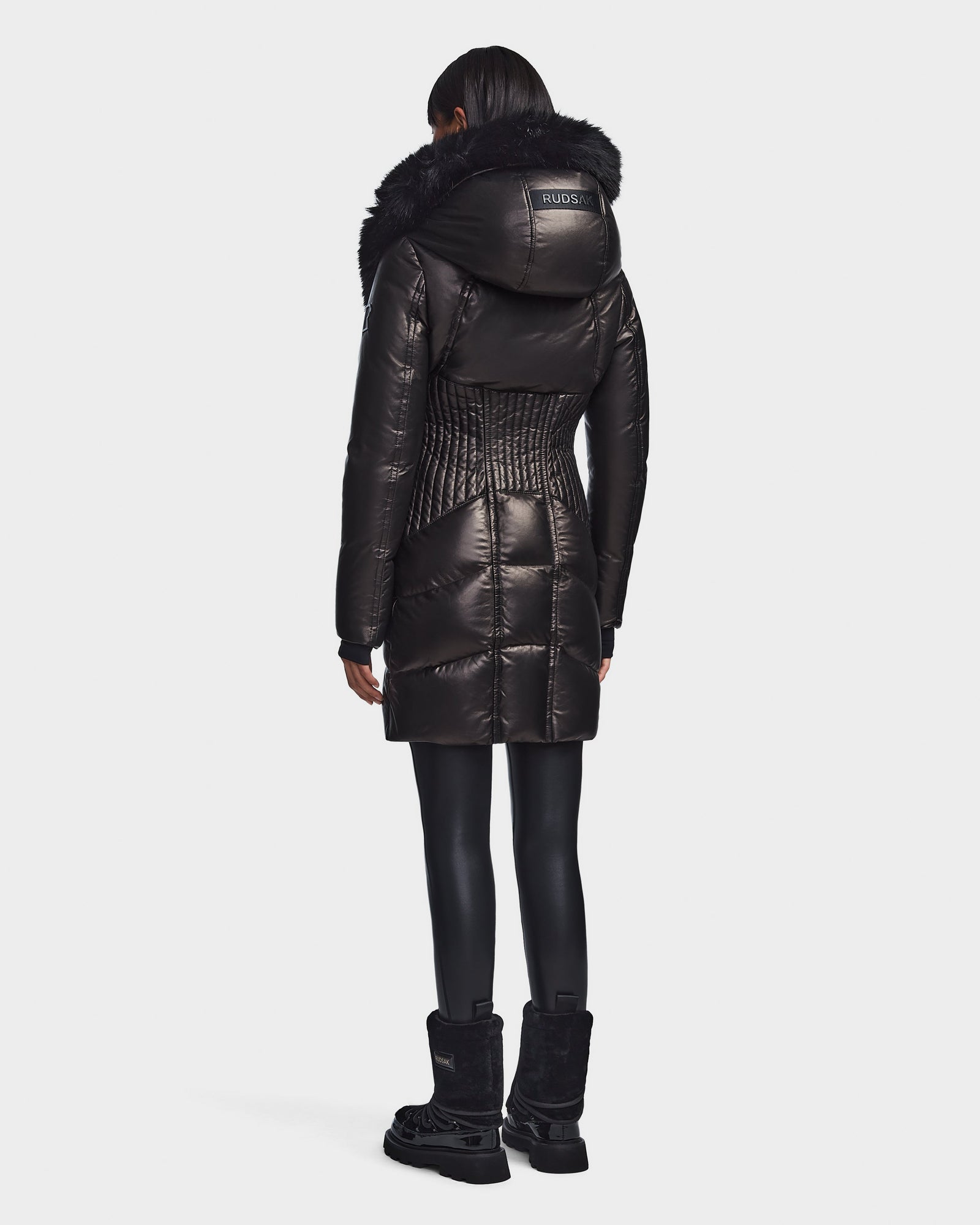 A person stands facing away, wearing the Rudsak SHAUNA X - 8121568 BLACK—a shiny black puffer with a large fur-trimmed hood—paired with black leggings and boots against a white background; a chic women's winter coat for cold days.