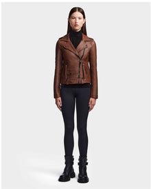 A woman stands facing forward in the brown RUDSAK MERGO - 8122009 COHIBA lambskin leather jacket, black turtleneck, leggings, and boots against a light gray background.