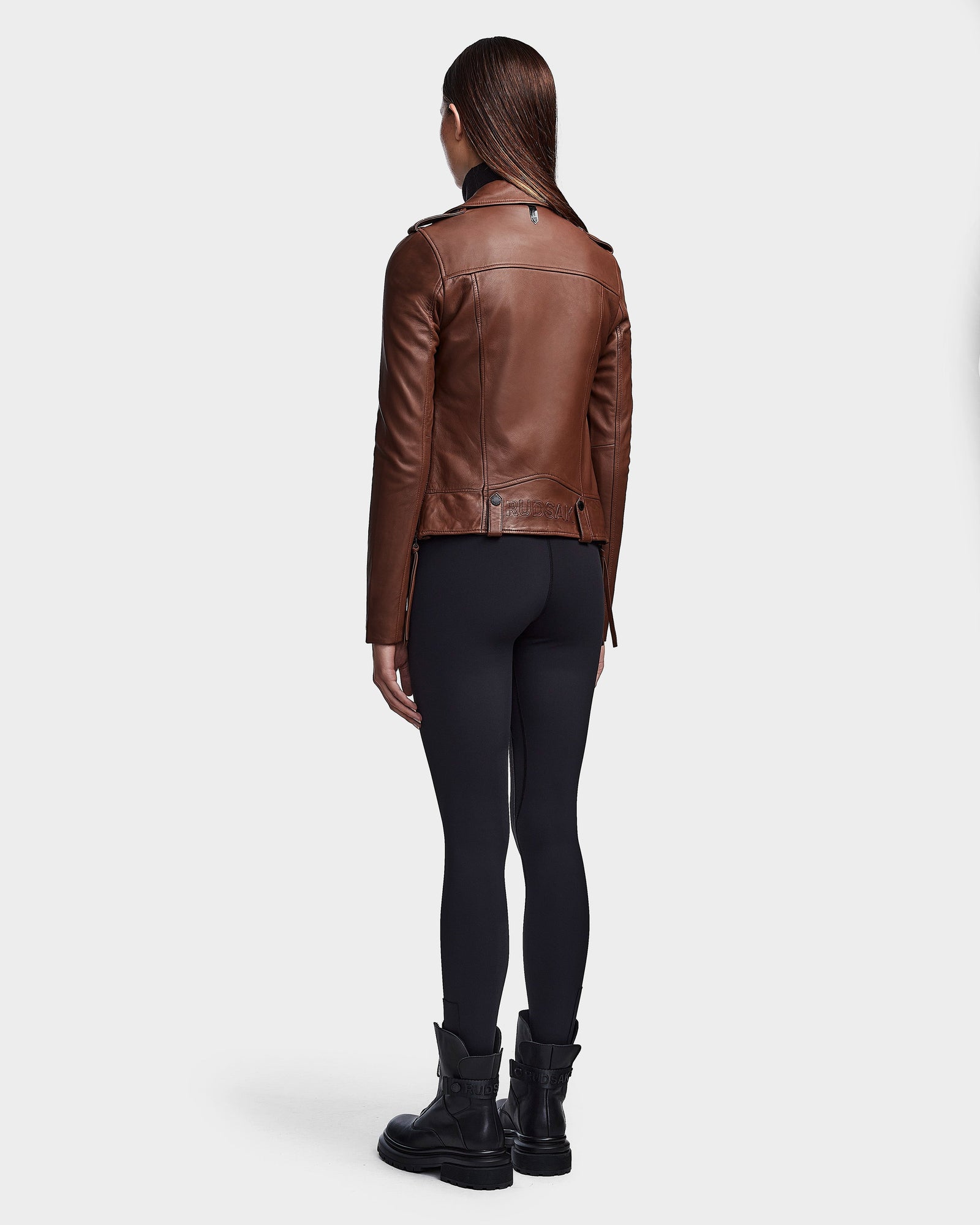 A person with straight, dark hair stands facing away, wearing the RUDSAK MERGO - 8122009 COHIBA women's brown lambskin leather biker jacket, paired with black leggings and boots against a plain light background.
